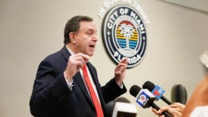 A man in a suit gestures passionately while speaking at a press conference behind the City of Miami seal, with microphones gathered around.