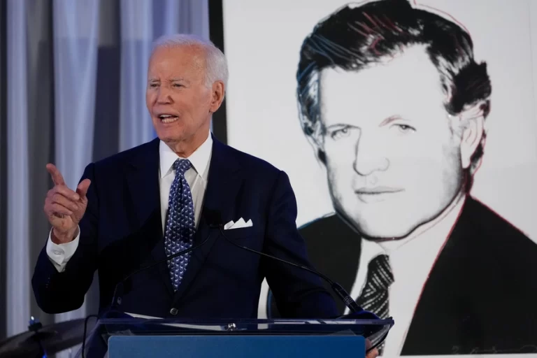 A man in a dark suit gestures while speaking at a podium, with a black and white portrait displayed behind him.