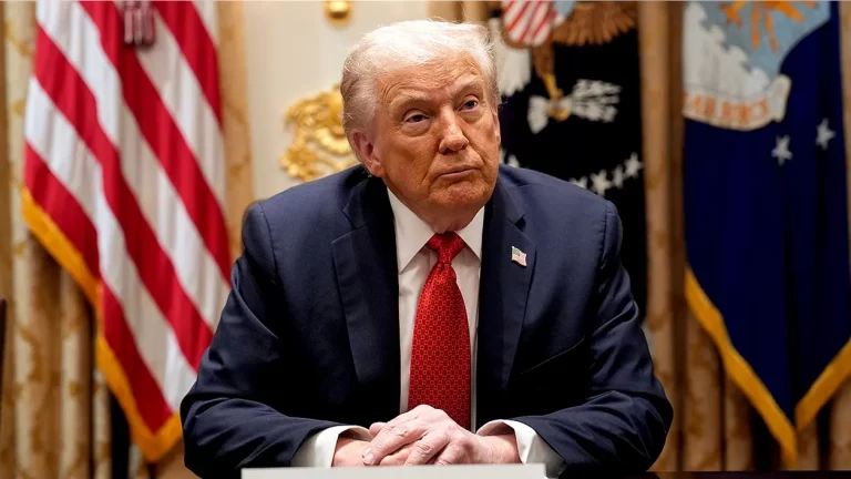 Man in navy suit and red tie sitting at desk in government office with U.S. flags in background, formal political setting, serious expression