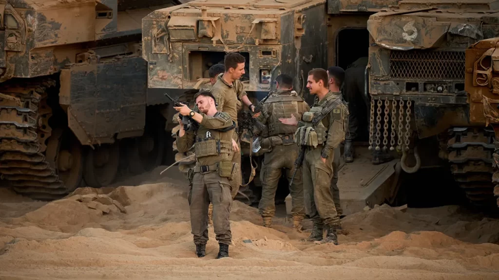 A group of soldiers in military gear stands in a sandy environment near armored vehicles
