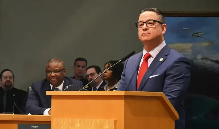 A man in a blue suit and red tie stands confidently at a podium speaking, while seated individuals listen attentively.