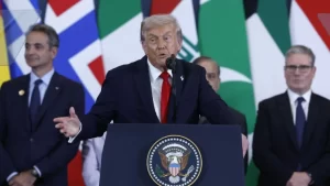 A man stands behind a podium adorned with the presidential seal, gesturing during a speech at an international event with flags in the background.