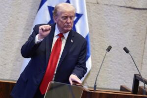 A man in a suit and red tie gestures while speaking at a podium, with an Israeli flag in the background.