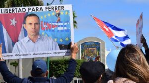 Protesters hold signs demanding freedom for Ferrer, with a Cuban flag waving in the background and a vibrant mural visible.