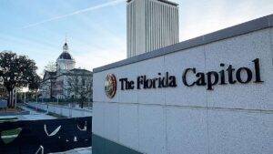 The Florida Capitol sign in the foreground with the historic Capitol building visible in the background under a clear blue sky.