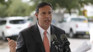 A man in a suit gestures while speaking at a microphone, with parked vehicles and trees blurred in the background.
