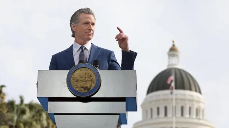 A politician speaks at a podium adorned with the California state seal, with a historic building in the background