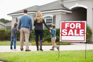 A family of four stands in front of a house for sale, holding hands and looking towards the home, with a prominent "For Sale" sign in view.