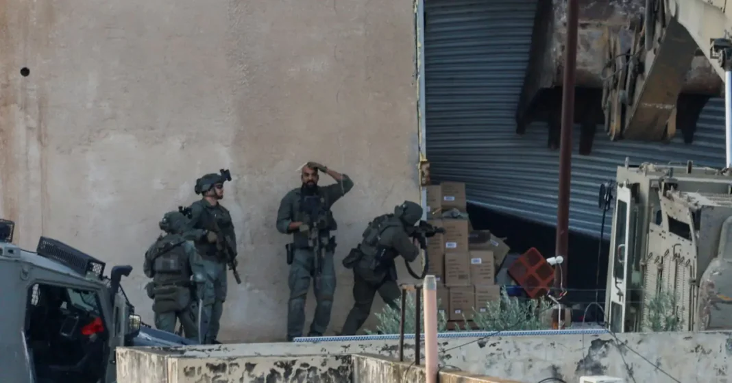 israel-west-bank-shooting-2025 Four armed soldiers in tactical gear stand near a wall beside a partially open warehouse door.