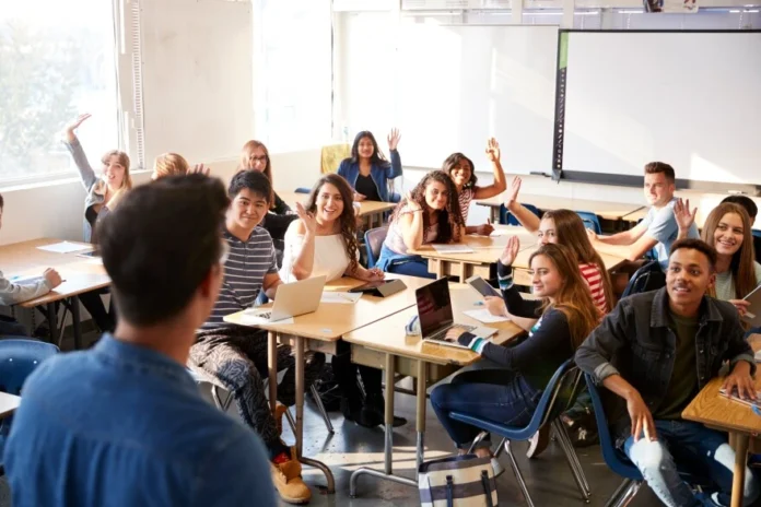 A classroom filled with students, many raising their hands, engaged in discussion while a teacher leads from the front.