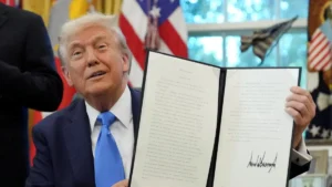 A person holds up a signed document in the Oval Office, with flags and a window in the background, indicating a formal announcement or signing event.