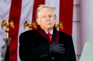 A man in a black coat and gloves stands with his hand over his chest in front of a large American flag and golden eagle decorations.