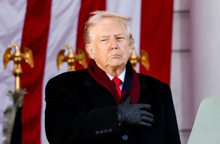 A man in a black coat and gloves stands with his hand over his chest in front of a large American flag and golden eagle decorations.