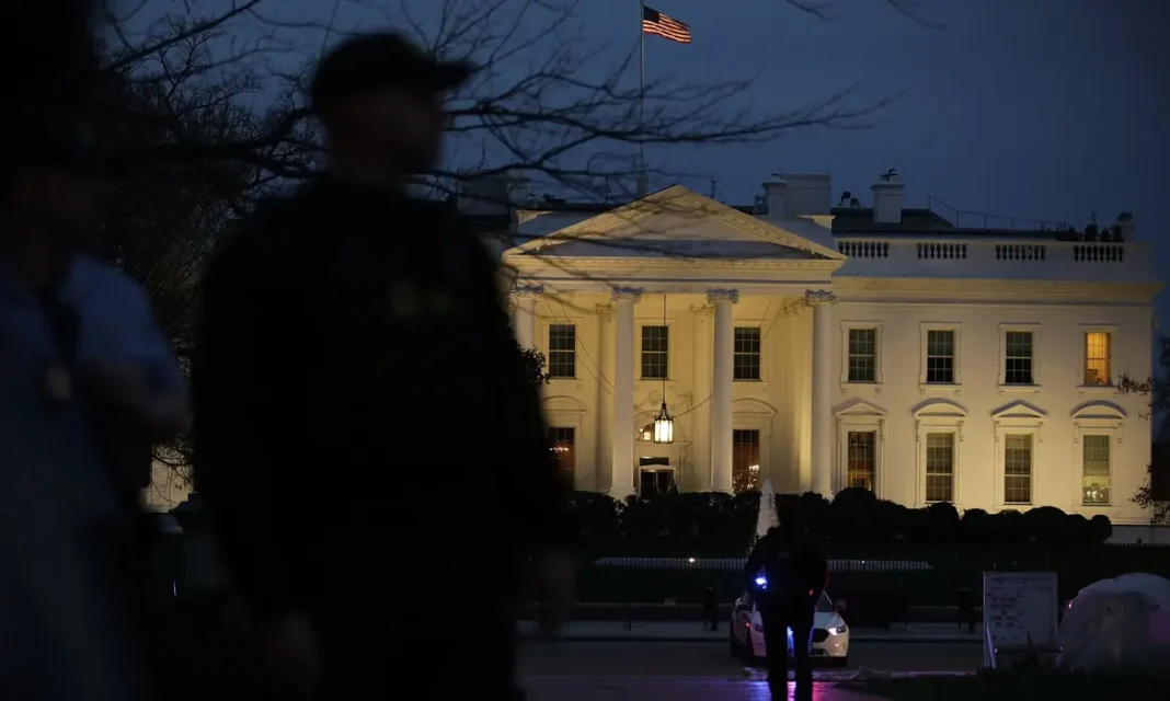 Trump Tightens U.S. Entry After Deadly D.C. Attack The White House illuminated at night, with silhouetted security personnel in the foreground and an American flag flying above.