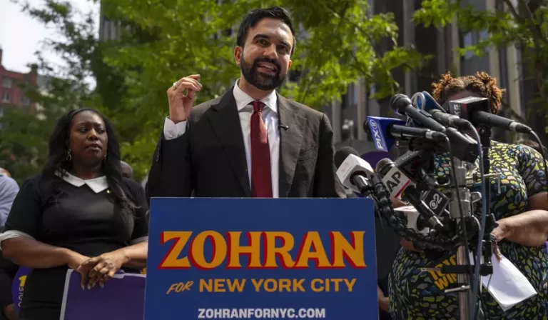 A man in a suit gestures while speaking at a podium reading "ZOHRAN for NEW YORK CITY," surrounded by press and supporters.