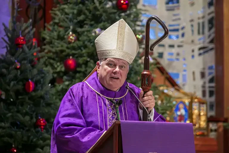 clergy member in purple robes and a white mitre speaks at a festive altar, decorated with Christmas trees and ornaments.