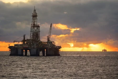 An offshore oil rig silhouetted against a dramatic sunset, with dark clouds and shimmering water surrounding it.