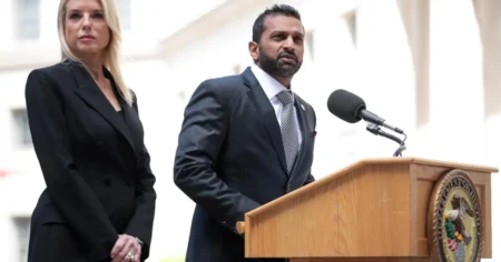A man in a suit speaks at a podium with the Department of Justice seal, while a woman stands beside him in a black outfit.