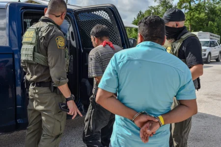A police officer assists a detained individual entering a police vehicle, while two men stand nearby, observing the situation.