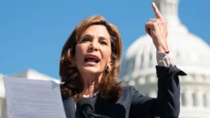 A woman speaks at a podium outdoors, holding papers in one hand and gesturing upward with the other, with a domed government building in the background.