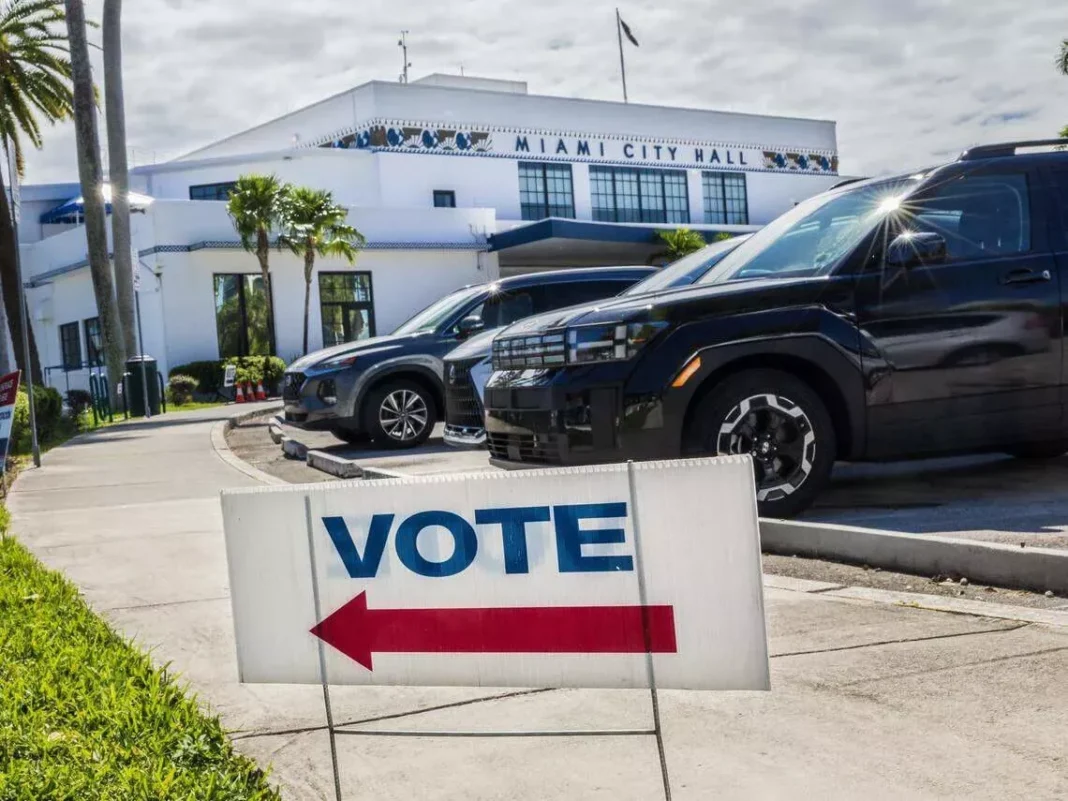 miami-city-hall-elections Vote sign with red arrow on sidewalk points towards Miami City Hall. Cars parked nearby, sunny day with palm trees.