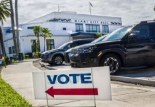 Miami-Dade Runoff Election: Voters in Three Cities Head to the Polls Today Vote sign with red arrow on sidewalk points towards Miami City Hall. Cars parked nearby, sunny day with palm trees.