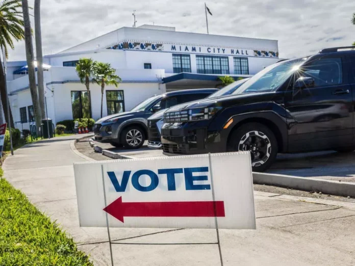 Vote sign with red arrow on sidewalk points towards Miami City Hall. Cars parked nearby, sunny day with palm trees.