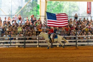 A rider on a white horse carries a large American flag in an indoor arena. A cheering crowd in the stands captures the event