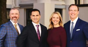group of four well-dressed individuals standing together outdoors, smiling, with a building and trees in the background.