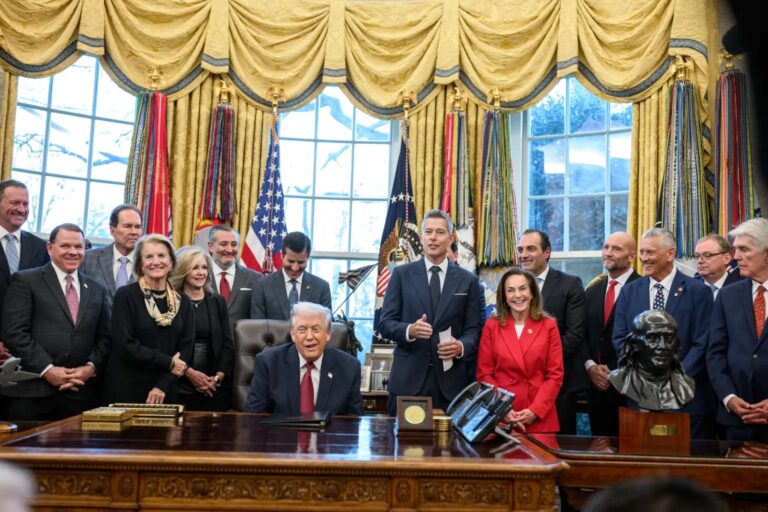 Trump Holds Economy-Focused Roundtable at the White House A group of officials, in formal attire, gathered around a desk in an ornate room, with American flags and curtains in the background.
