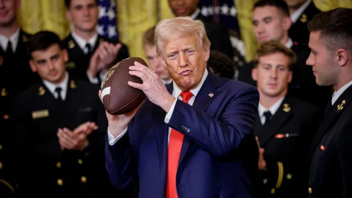 A man in a suit holds a football, flanked by applauding individuals in military uniforms, against a backdrop of American flags.