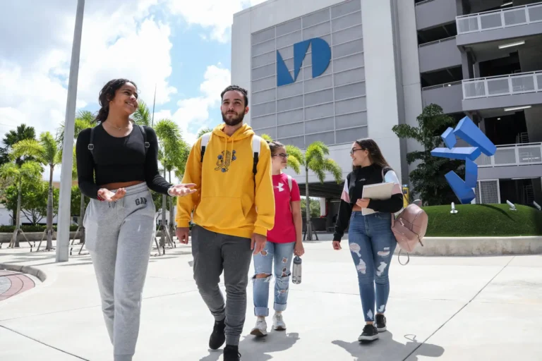 Four smiling students walk together outside a modern building with an "MDC" logo, surrounded by palm trees under a bright blue sky.