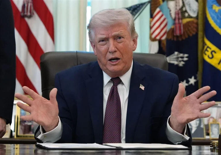 Person in a suit seated at a desk with hands raised, American flags and official symbols in the background.