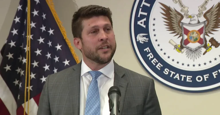 Man in suit speaking at a podium with an American flag and a state attorney seal in the background.