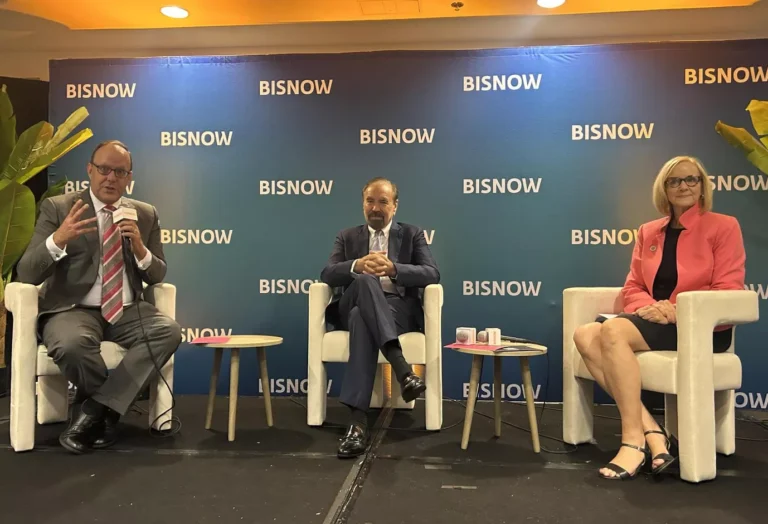 Three people in business attire seated on stage with "BISNOW" backdrop during a panel discussion.