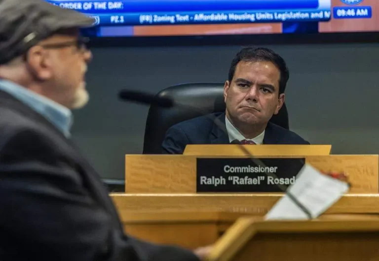 Two men engaged in a formal meeting, one seated behind a desk labeled "Commissioner Ralph 'Rafael' Rosado."