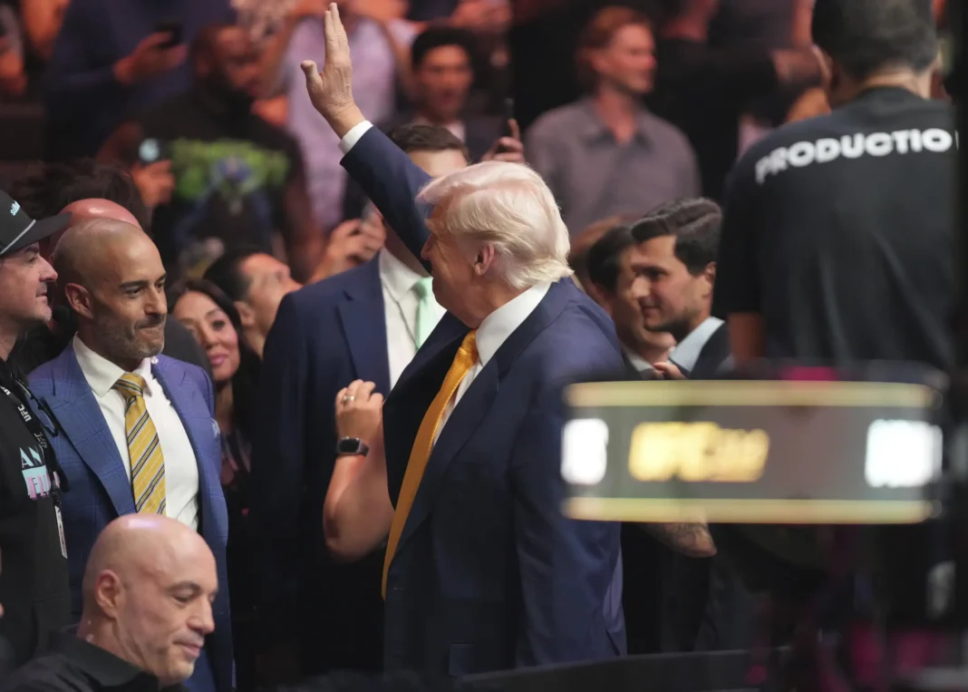 Man in a suit with a yellow tie raising his hand in a crowded indoor event with people and security around.