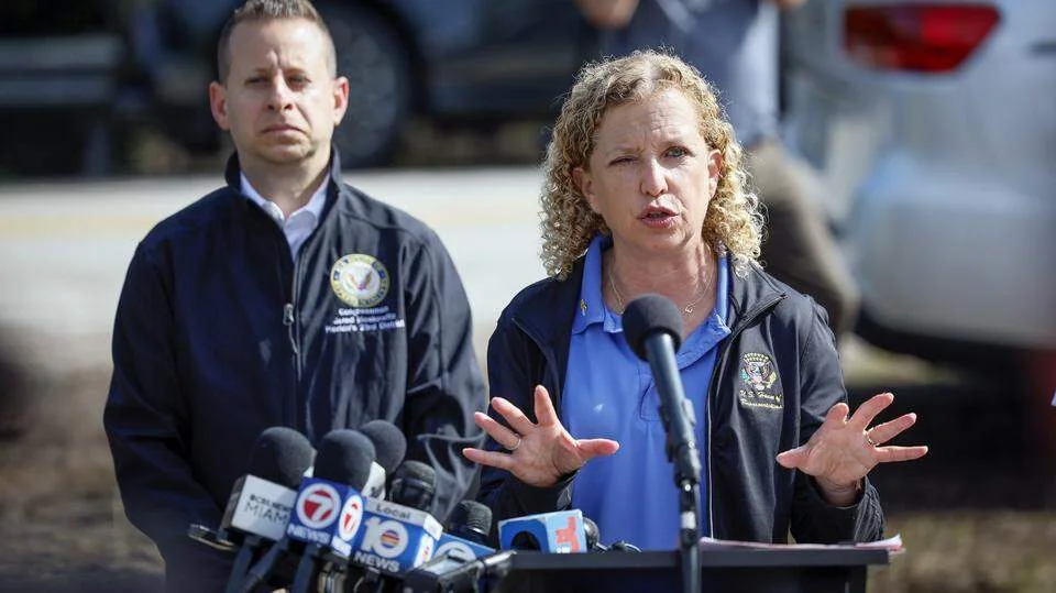 Two officials wearing jackets speak at a press conference with multiple microphones outdoors.