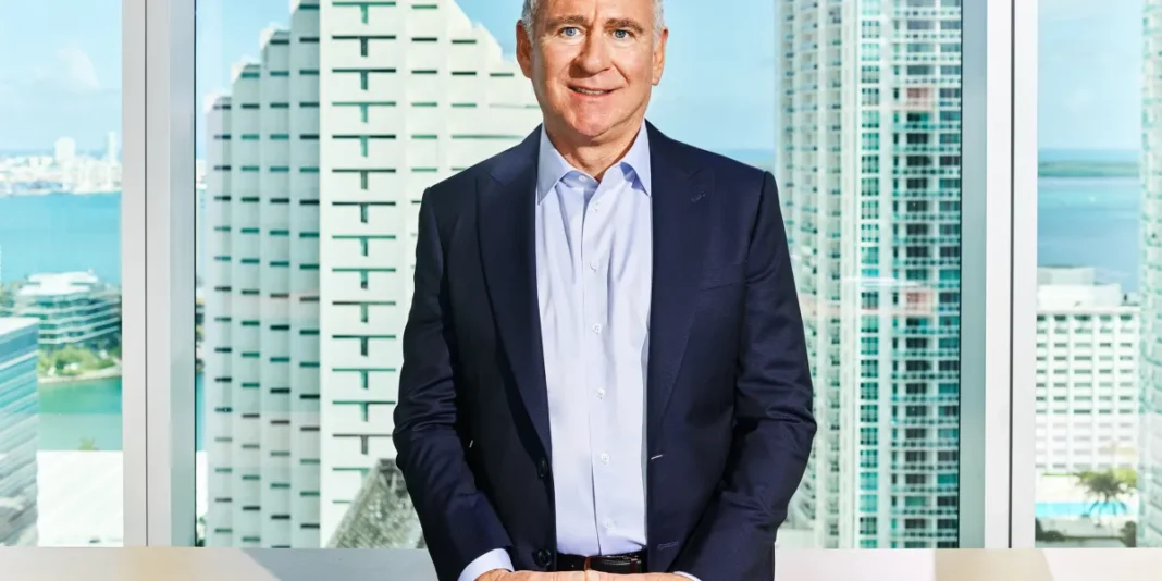 Middle-aged man in a navy suit stands smiling behind a desk with city buildings and water visible through large windows.