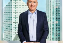 Ken Griffin Expands Miami Footprint as Businesses Flee High-Tax Policies Middle-aged man in a navy suit stands smiling behind a desk with city buildings and water visible through large windows.