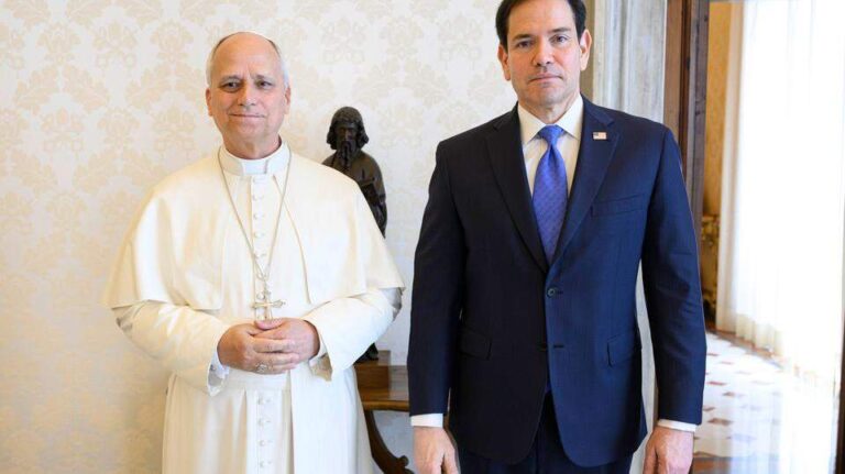Two men stand indoors against a floral wallpapered wall, one in white religious attire and the other in a dark suit with a blue tie.