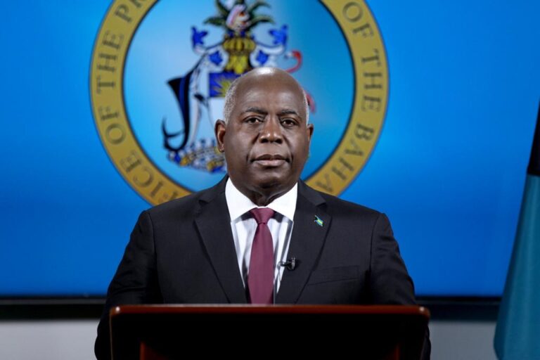 A man in a suit stands before a podium, a serious expression on his face. The Bahamian flag and official seal are visible in the blue background.