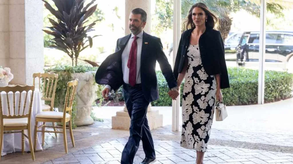 A man in a suit and a woman in a black and white floral dress holding hands, walking outdoors near chairs and plants.