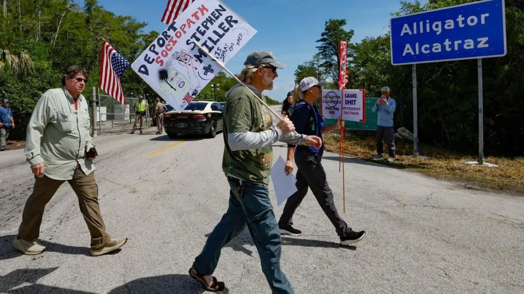 Florida Faces New Lawsuit Over “Alligator Alcatraz” Migrant Detention Center Protesters holding signs and American flags walk past a sign reading "Alligator Alcatraz" on a sunny day.