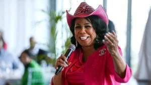 Person in a bright pink outfit and matching hat speaking into a microphone, gesturing with their hand in a blurred indoor setting.