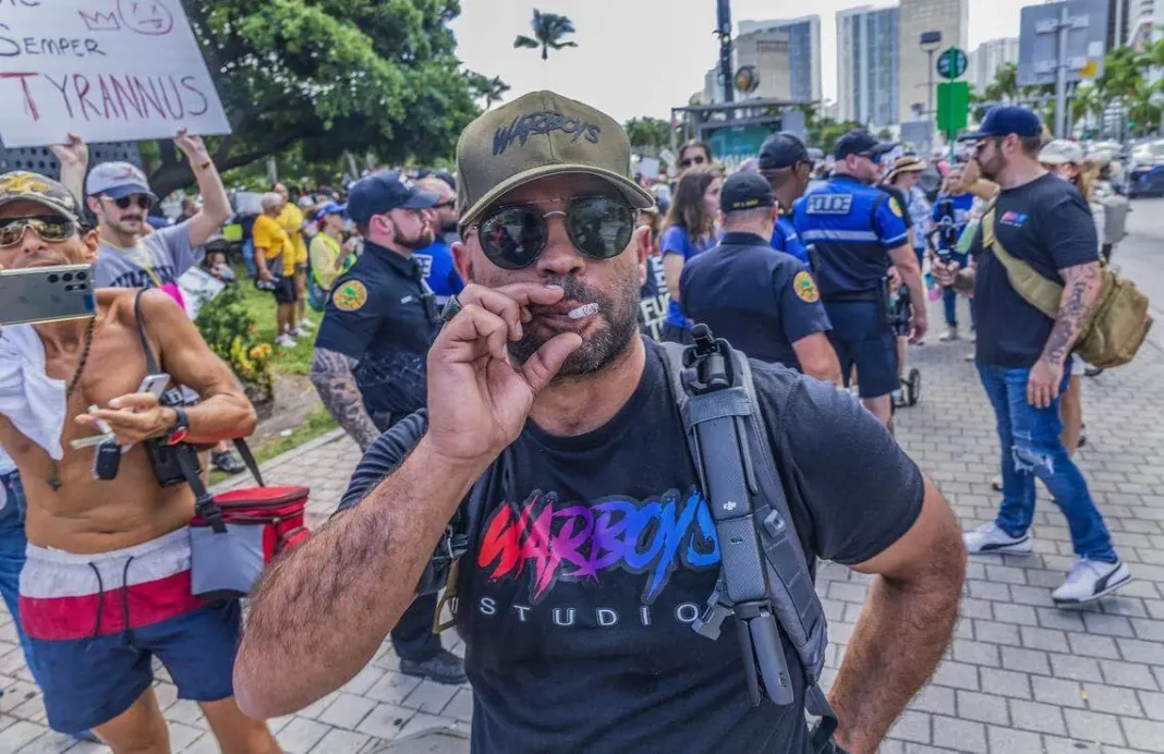 Proud Boys Leader Tarrio Expects Huge Trump Compensation Man in Garboys Studio shirt holding phone at a street protest with police and other demonstrators in the background.