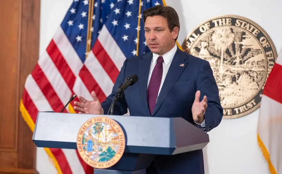 Florida Policy Institute Warns DeSantis Property Tax Plan Could Cost Billions Person in suit speaking at a podium with the Great Seal of Florida, with American and Florida state flags in the background.
