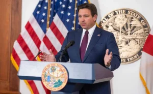 Person in suit speaking at a podium with the Great Seal of Florida, with American and Florida state flags in the background.