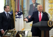 Trump Says He Is “In No Hurry” to Finalize Iran Deal as Nuclear Demands Remain Central A man in a suit speaks at a podium with the presidential seal, while another man in a suit stands nearby in a formal room with flags.