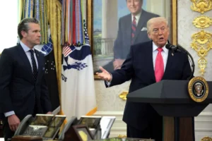 A man in a suit speaks at a podium with the presidential seal, while another man in a suit stands nearby in a formal room with flags.